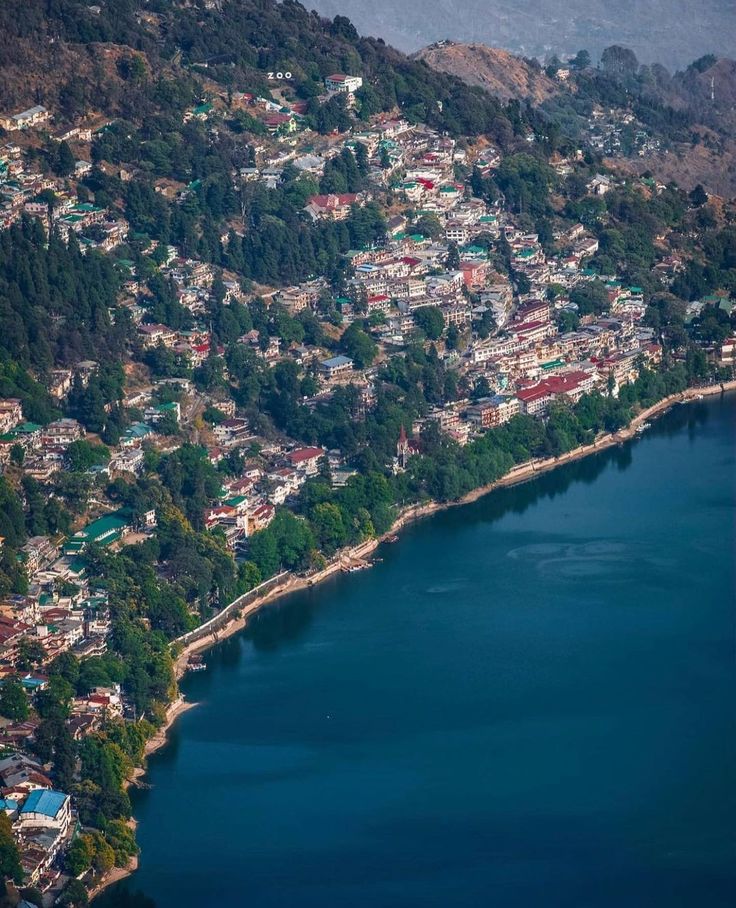 यात हे असू शकते: पार्श्वभूमीवर पर्वत असलेल्या तलावाच्या काठावर असलेल्या एका लहान शहराचे हवाई दृश्य