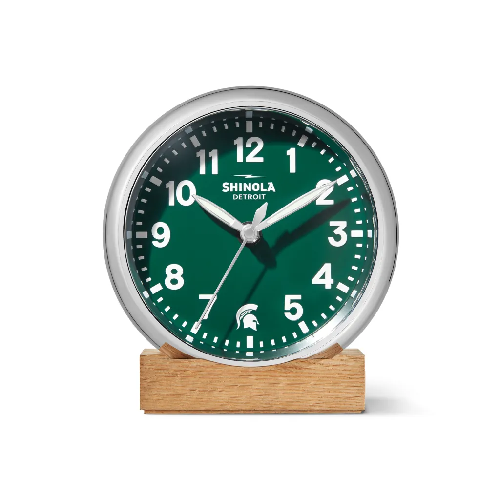 गडद हिरवा चेहरा, क्रोम ट्रिम, पांढरे अंक आणि मिशिगन स्टेट स्पार्टन्स हेल्मेट लोगो असलेले शिनोला डेट्रॉईट घड्याळ.