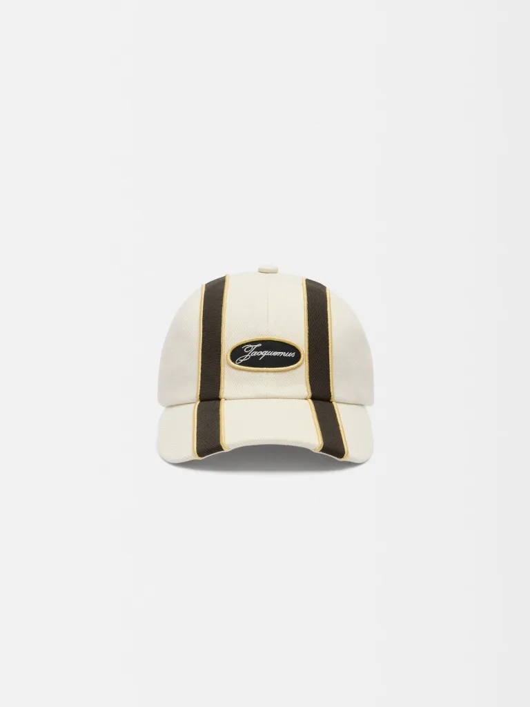काळ्या आणि सोन्याच्या पट्ट्यांसह क्रीममध्ये जॅक्युमस बेसबॉल कॅप.