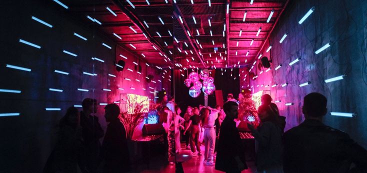 यात याचा समावेश असू शकतो: भिंती आणि मजल्यावरील निऑन दिवे असलेल्या बोगद्यात उभा असलेला लोकांचा गट