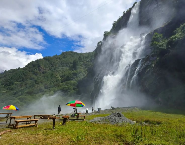 यात हे असू शकते: छत्री असलेले दोन लोक धबधब्यासमोर उभे आहेत आणि त्यांच्या खाली पिकनिक टेबल आहेत