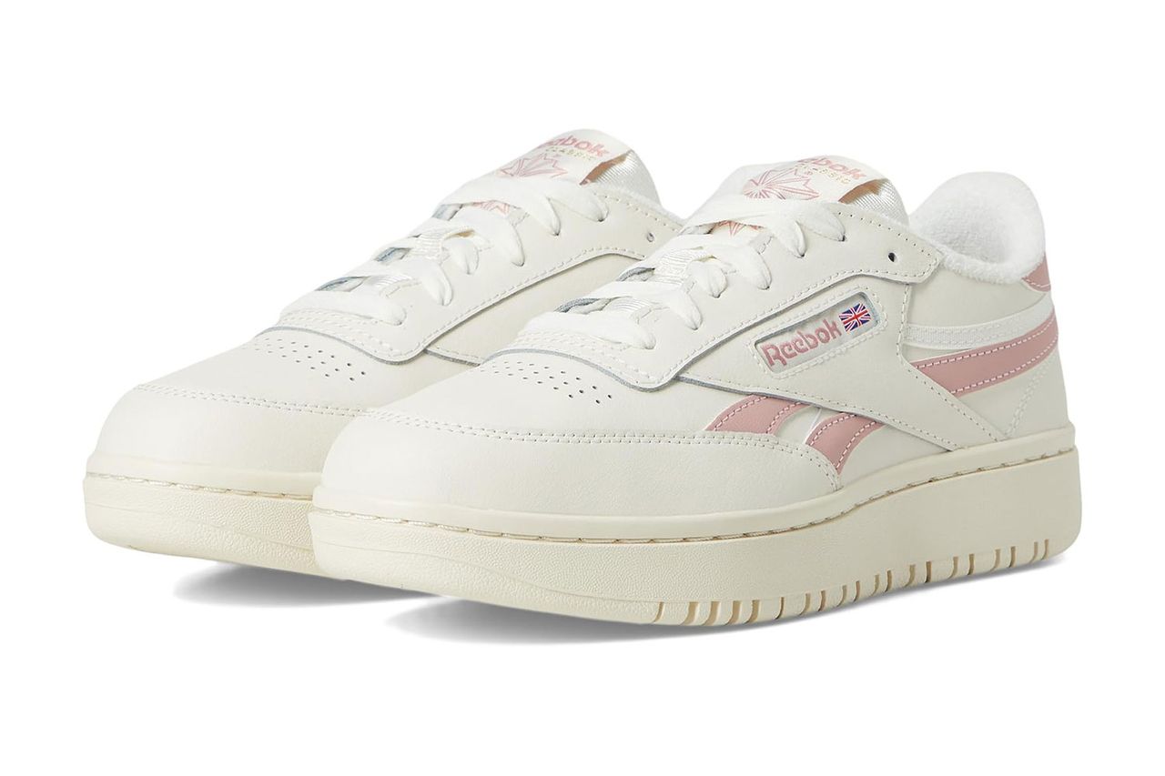 रीबॉक क्लब सी डबल स्नीकर्सची जोडी एका बाजूच्या पट्टीच्या तपशीलासह साध्या पार्श्वभूमीवर ठेवली आहे झाप्पोस