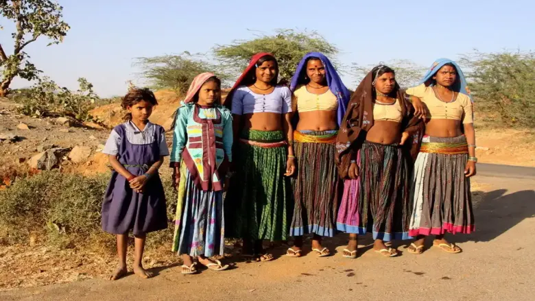 पारंपारिक रंगीबेरंगी घागरा-चोली आणि डोक्यावर स्कार्फ घालून राजस्थानमधील ग्रामीण रस्त्याच्या कडेला उभ्या असलेल्या गरसिया आदिवासी महिला आणि एक तरुण मुलगी. त्यांचा पोशाख आणि मुद्रा गारसिया समुदायाची सांस्कृतिक ओळख, दैनंदिन जीवन आणि पारंपारिक जीवनशैली प्रतिबिंबित करतात.