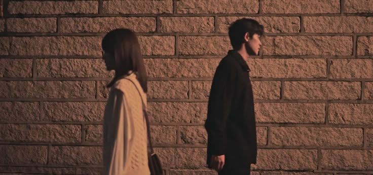 यात हे असू शकते: वीट भिंतीसमोर दोन लोक उभे आहेत आणि एक व्यक्ती दुसऱ्याकडे पाहत आहे