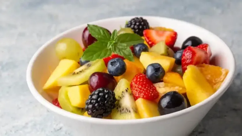 स्ट्रॉबेरी, ब्लूबेरी, ब्लॅकबेरी, किवी, आंब्याचे तुकडे आणि हिरवी द्राक्षे असलेल्या ताज्या फळांच्या सॅलडने भरलेला एक चमकदार पांढरा वाडगा, पुदिन्याच्या ताज्या कोंबांनी भरलेला.