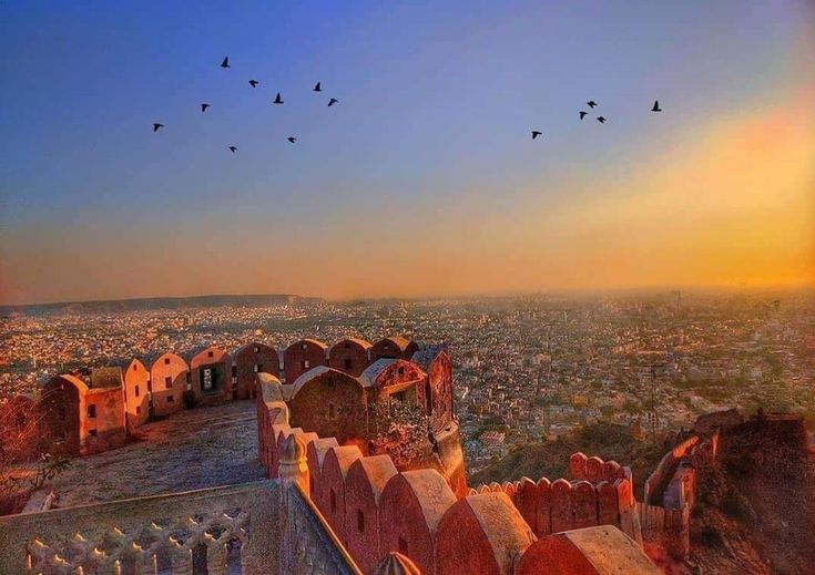 यात हे असू शकते: पक्षी सूर्यास्ताच्या किंवा सूर्योदयाच्या वेळी जुन्या इमारतीच्या छतावरून उडत आहेत