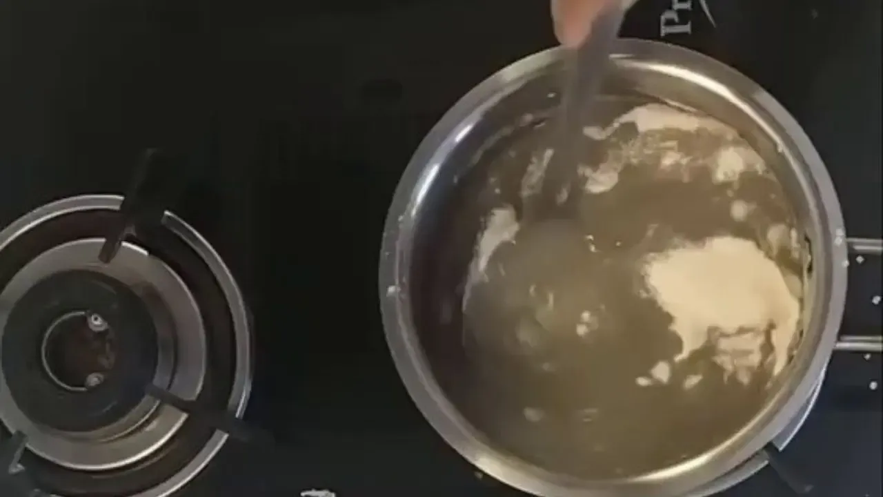 एका हाताने काळ्या इंडक्शन स्टोव्हवर स्टेनलेस स्टीलच्या भांड्यात उकळत्या पाण्यात पांढरे पीठ ढवळले.