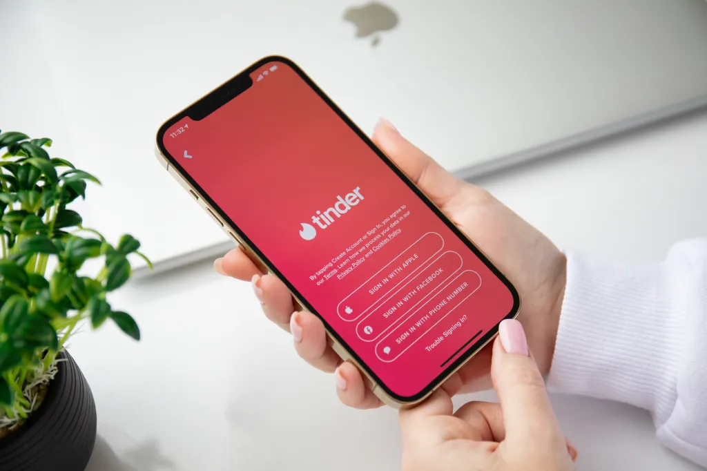 सोन्याचा iPhone 12 Pro Max हातात धरलेली स्त्री Tinder ॲप साइन-इन स्क्रीन दाखवत आहे.